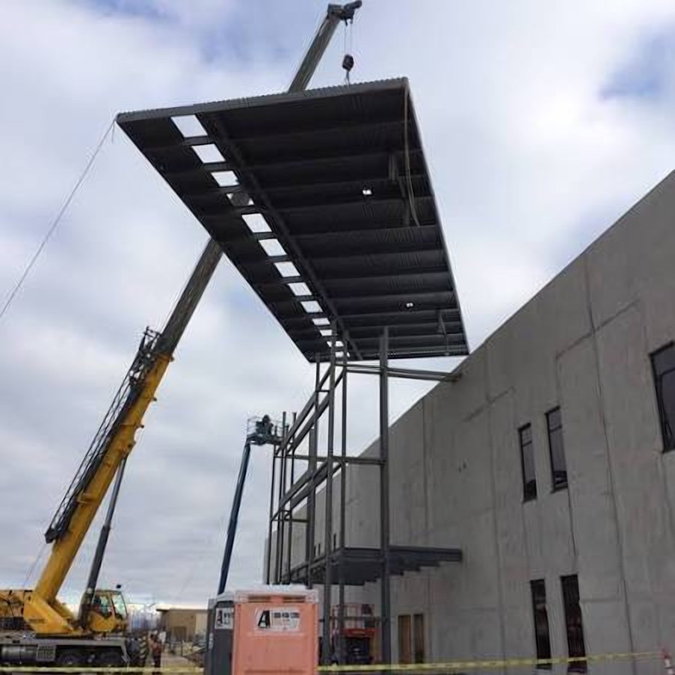 A crane lifting a steel canopy frame into position beside a concrete building