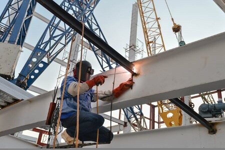 A welder working on a steel beam at height beside a crane